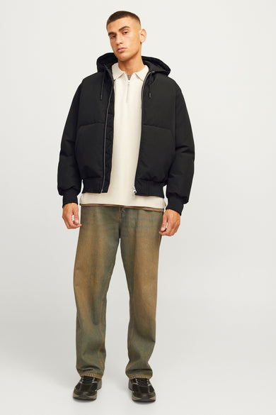 Zwarte korte jas van CORE BY JACK & JONES, met capuchon en gedragen over een beige shirt en groen-bruine jeans.
