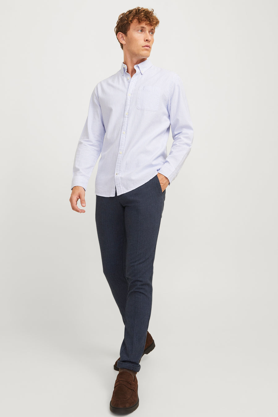 Man in gestreept overhemd, donkere broek en bruine schoenen op witte achtergrond.