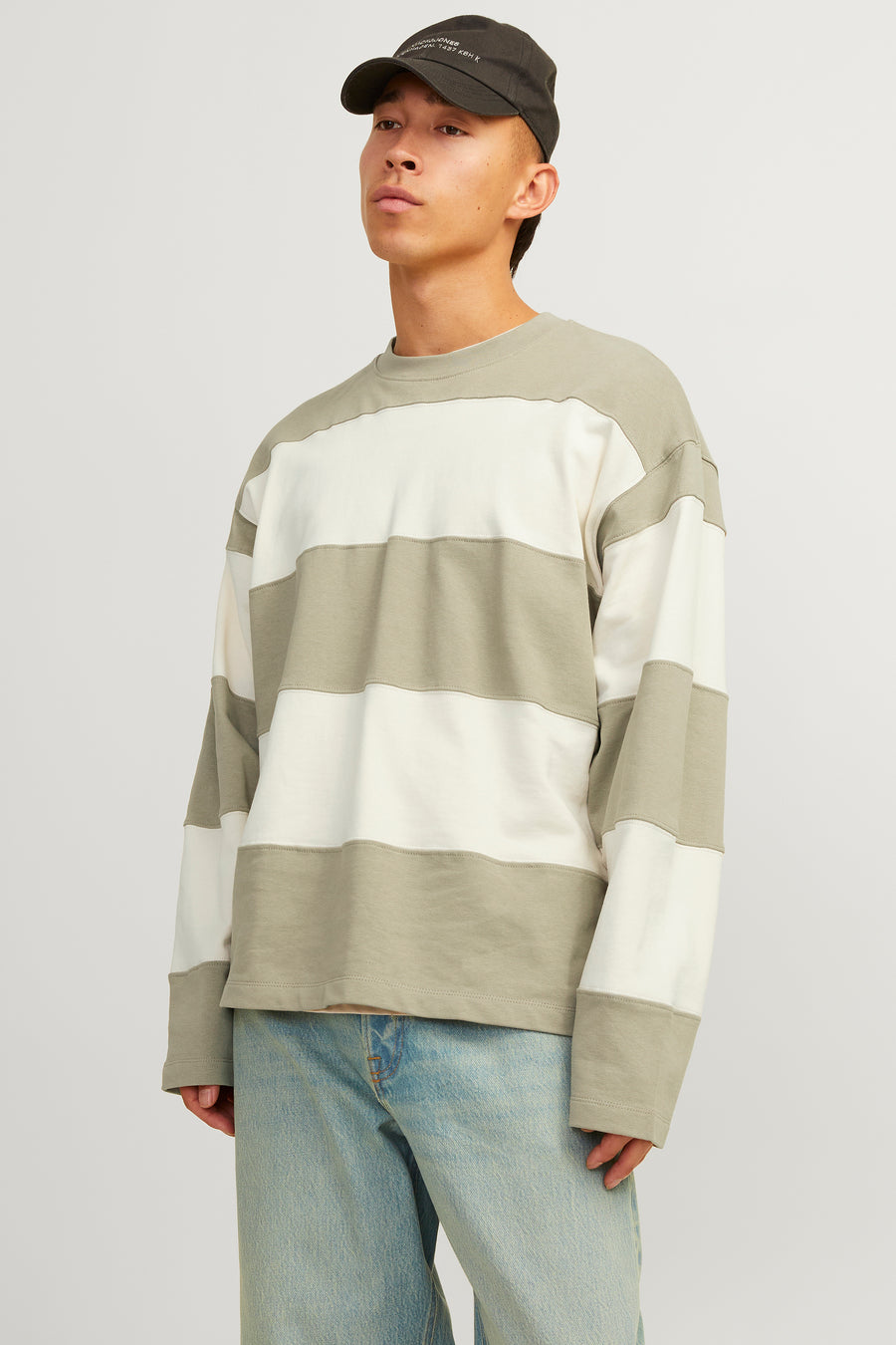 Een man draagt een gestreepte trui en jeans, met een pet op, staand tegen een witte achtergrond.