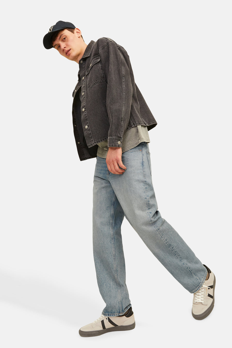 Een man in een zwarte spijkerjas, blauwe jeans, sneakers en een pet.