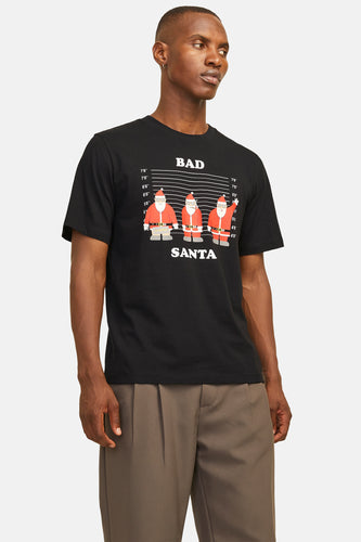 Een man met een zwart T-shirt met 'Bad Santa'-print en bruine pantalon tegen een neutrale achtergrond.