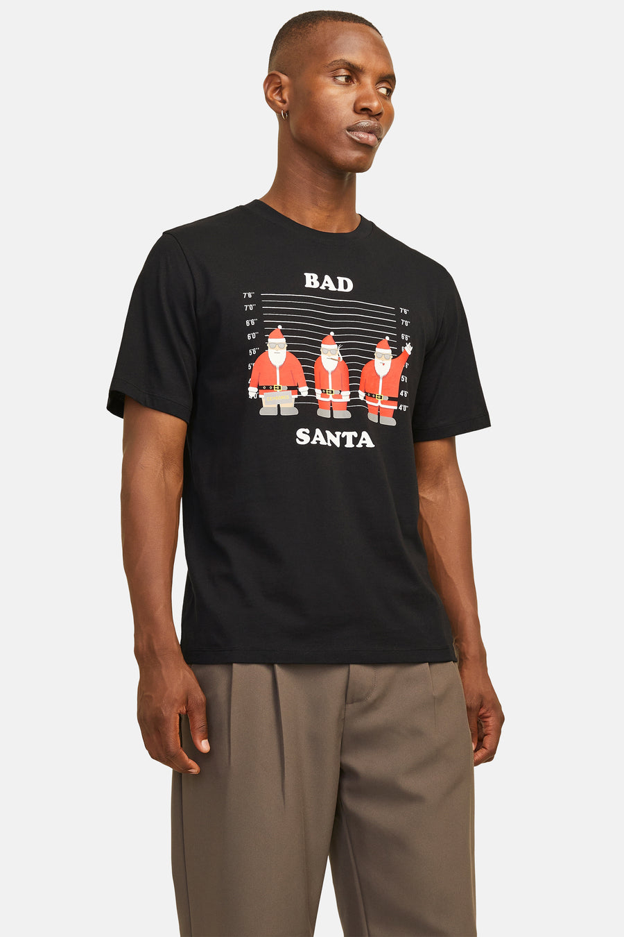 Een man met een zwart T-shirt met 'Bad Santa'-print en bruine pantalon tegen een neutrale achtergrond.