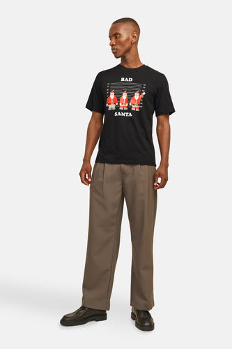 Man met zwart T-shirt met "Bad Santa"-opdruk, bruine pantalon en zwarte schoenen, staand tegen een witte achtergrond.