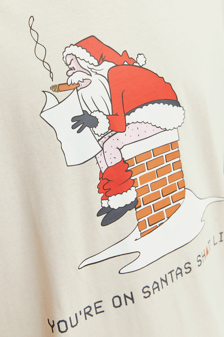 Beige T-shirt met een cartoon van de Kerstman die een sigaar rookt en uit een schoorsteen komt, met de tekst "You're on Santa's Sh*t List".