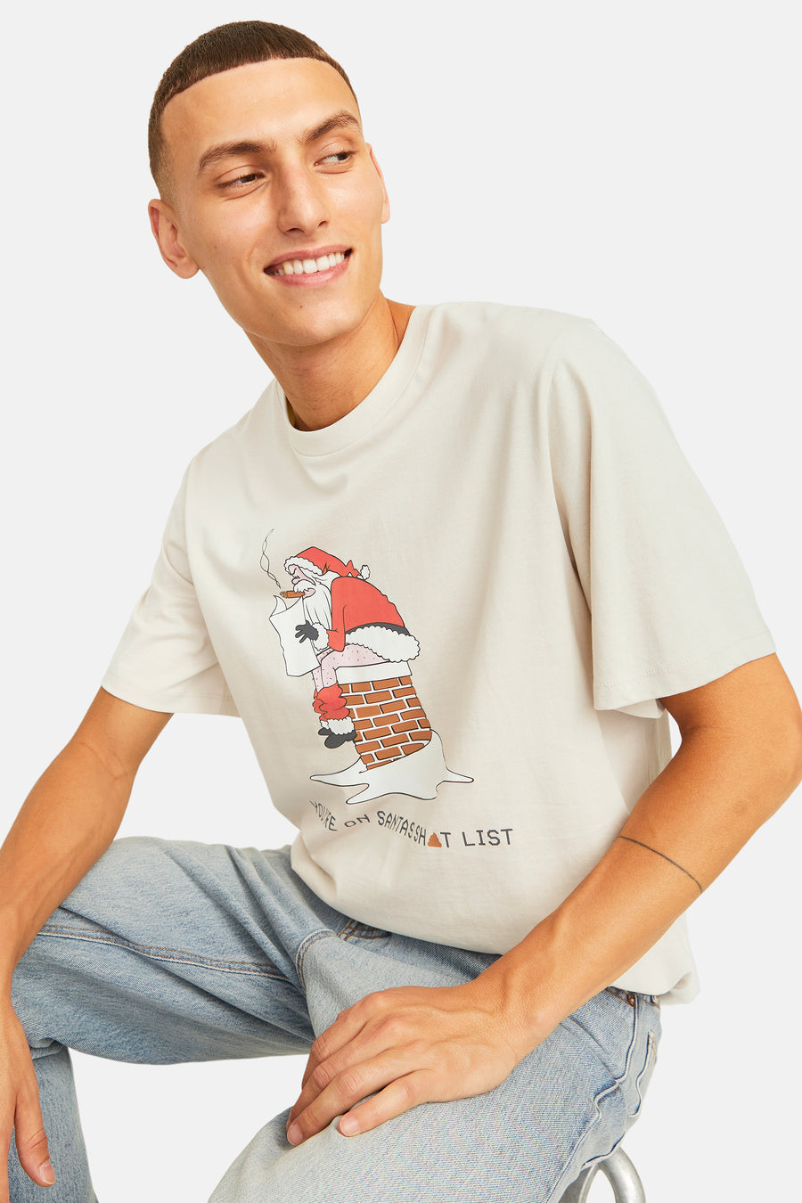 Man met beige T-shirt met Santa print, zittend in jeans.
