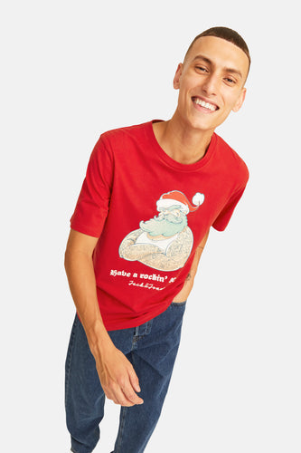 Lachende man met rood T-shirt met kerstmotief en blauwe jeans, staand op witte achtergrond.