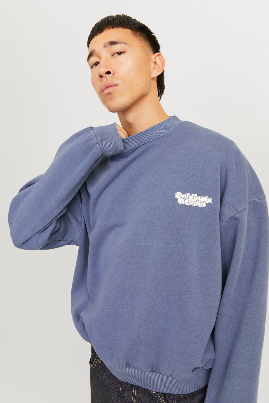 Man met blauwe trui met 'Originals Studio'-opdruk, staande tegen een witte achtergrond.