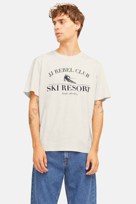 T-shirt beige à manches courtes de JJREBEL, avec une impression d'un resort de ski et porté avec un jean bleu.