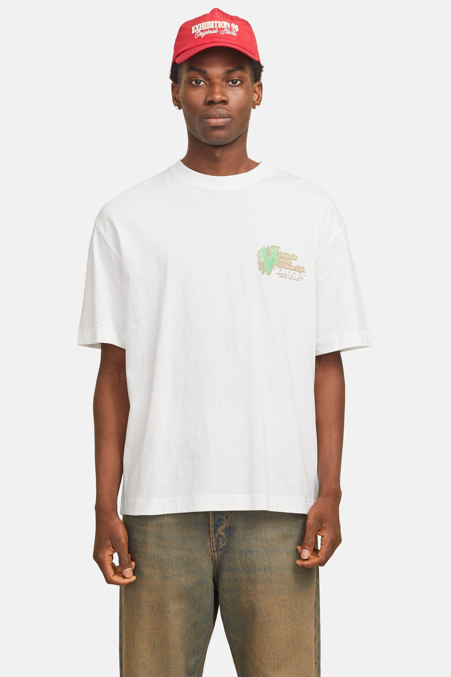 Een man in een witte T-shirt met een print en een rode pet, staand voor een effen achtergrond.