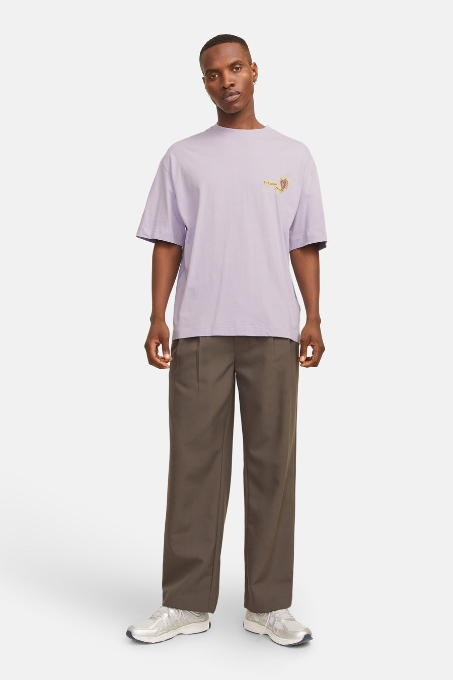 Een man staat in een lila shirt en bruine broek, met zilveren schoenen op een witte achtergrond.