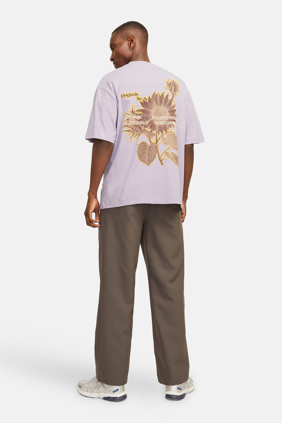 Man van achteren, lila T-shirt met zonnebloemprint, bruine broek, witte sneakers.