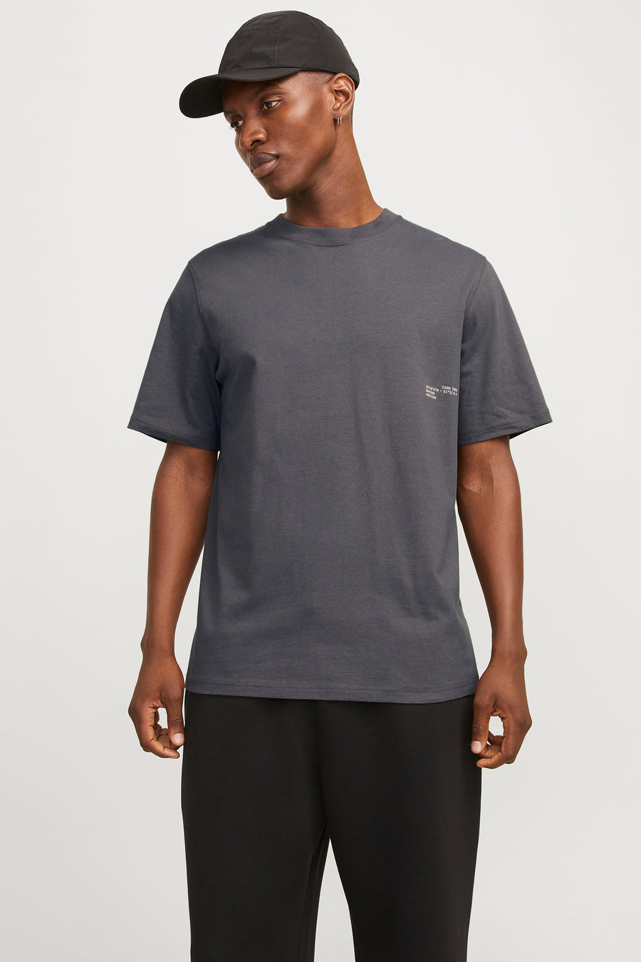 Man in grijs T-shirt en zwarte pet, staand tegen witte achtergrond.