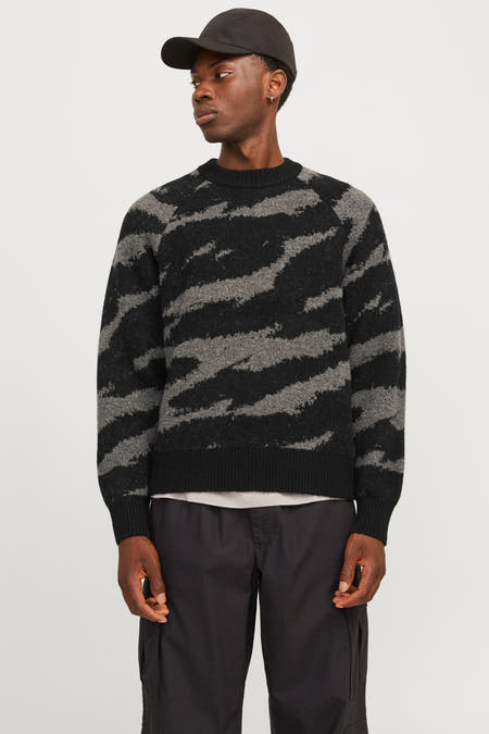 Un pull noir à col rond de CORE BY JACK & JONES, avec un motif gris ondulé, porté sous un bomberjack noir, associé à des pantalons cargo noirs et des bottes brunes.