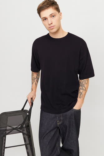 T-shirt noir à manches courtes de PREMIUM by JACK & JONES, porté avec des jeans foncés et assis sur une chaise en métal.
