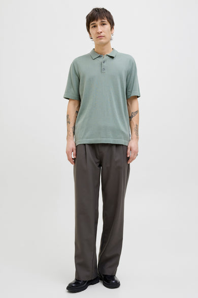 Man met tattoo's, groene polo, grijze pantalon en zwarte schoenen, staand voor witte achtergrond.