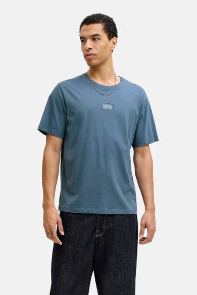 T-shirt bleu à manches courtes de CORE by JACK & JONES, avec un logo sur la poitrine, associé à des jeans foncés et des chaussures marrons.
