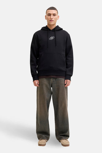 Man met zwarte hoodie, ruime jeans en suède mocassins op witte achtergrond.
