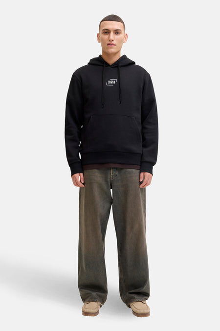 Man met zwarte hoodie, ruime jeans en suède mocassins op witte achtergrond.
