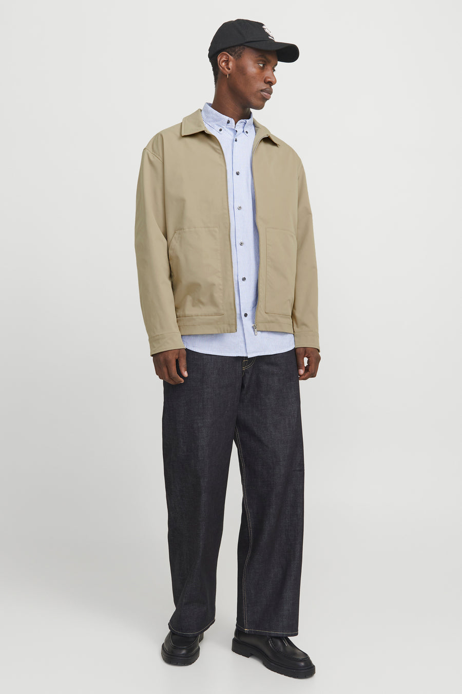 Man met pet, beige jas, gestreept overhemd en donkere jeans.