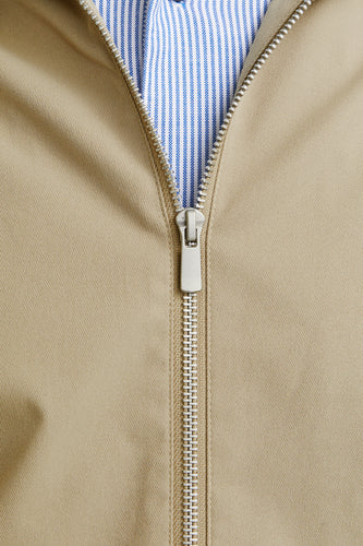 Beige jas met zilveren ritssluiting en blauw gestreept overhemd.