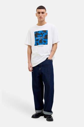 T-shirt blanc à manches courtes de CORE BY JACK & JONES, avec un motif bleu et noir sur la poitrine, porté avec des jeans bleus foncés et des chaussures noires.
