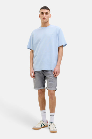 Jean court en denim gris de JACK & JONES JEANS INTELLIGENCE, avec des détails froissés et des poches repliées, porté avec un T-shirt bleu clair et des chaussettes blanches.