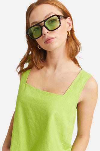 Vrouw met groene top, zonnebril en gouden oorbellen, witte achtergrond.