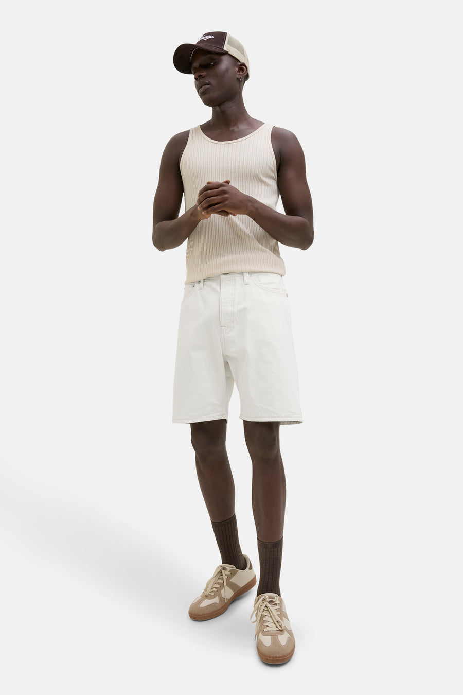 Man met bruine pet, mouwloze top, witte shorts, bruine sokken en beige sneakers op witte achtergrond.