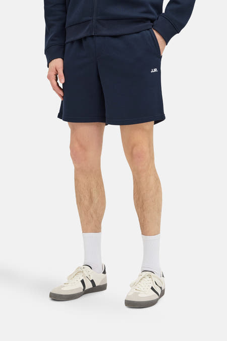 Short bleu marine de JJREBEL, avec impression de logo blanc, porté avec des chaussettes blanches et des baskets couleur crème.