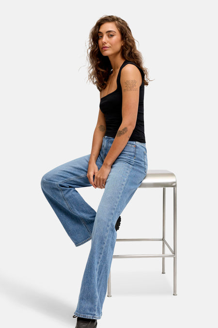 Jean évasé bleu moyen de JJXX, porté avec un haut noir, assis sur un tabouret en métal.