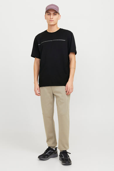 T-shirt noir à manches courtes de CORE par JACK & JONES, avec une bande horizontale sur la poitrine, associé à un pantalon de jogging beige et des baskets noires.