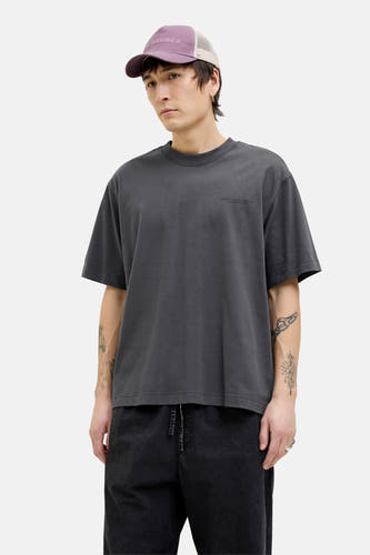 T-shirt gris à manches courtes de CORE BY JACK & JONES, avec texte sur la poitrine, porté avec une casquette et un pantalon noir.