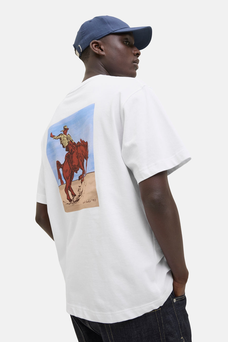T-shirt blanc à manches courtes d'ORIGINALS BY JACK & JONES, avec un imprimé cowboy sur le dos, associé à une casquette bleu marine et des jeans.