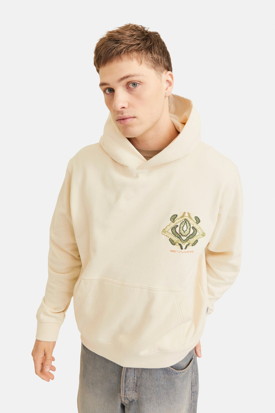 Een man draagt een crèmekleurige hoodie met een geborduurd logo en jeans tegen een witte achtergrond.