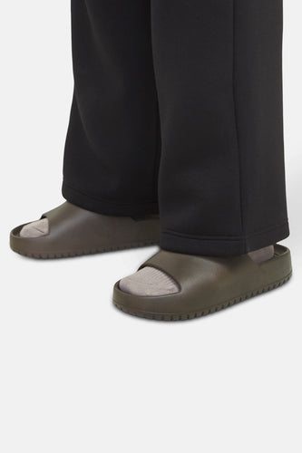 Persoon met zwarte joggingbroek en kaki sandalen met sokken, staand op een witte achtergrond.