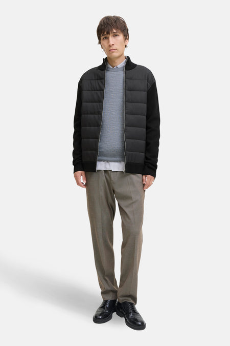 Man in gewatteerd bomberjack, grijze broek en zwarte schoenen, staand.