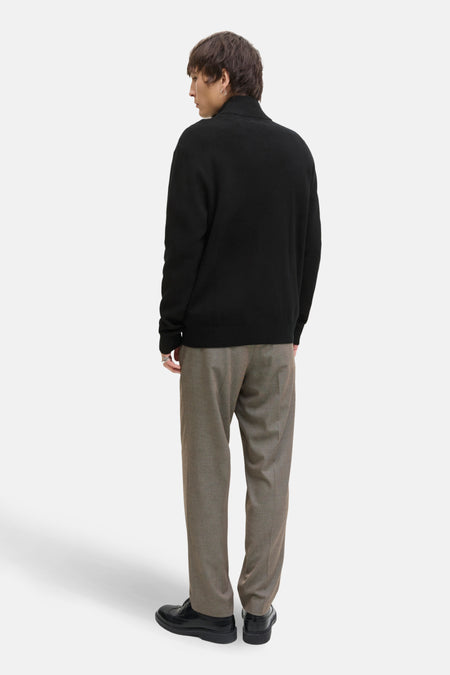 Man met zwarte trui, bruine broek en zwarte schoenen, van achteren gezien, witte achtergrond.