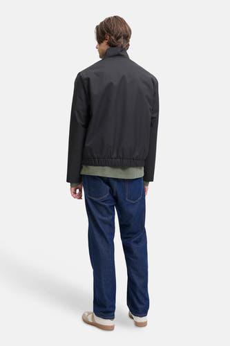 Manteau court noir de JJREBEL, vu de derrière, associé à un t-shirt vert olive, des jeans noirs et des chaussures crème.