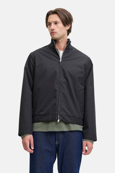 Manteau court noir de JJREBEL, avec des manches longues et une fermeture éclair argentée, porté sur un t-shirt vert olive et des jeans bleu marine.