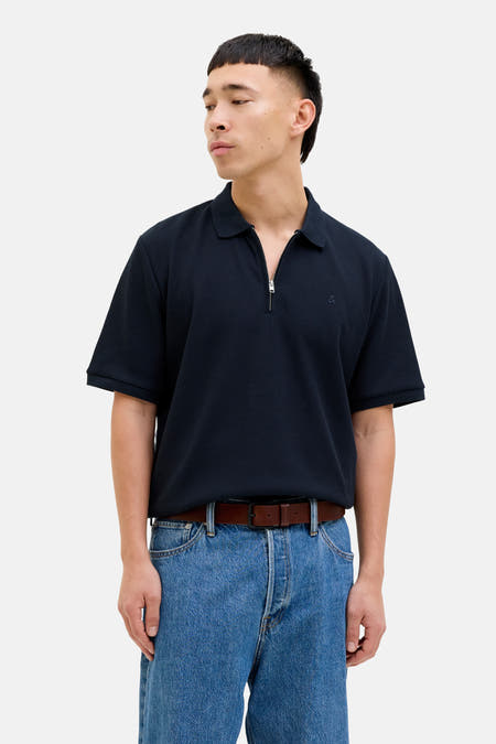 Polo bleu de PREMIUM par JACK & JONES, avec une fermeture éclair et porté avec un ceinture marron et des jeans.