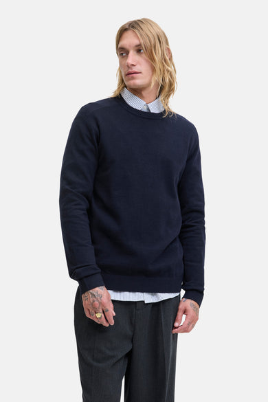 T-shirt bleu avec col rond de PREMIUM by JACK & JONES, porté sur un chemisier rayé et avec un pantalon gris.