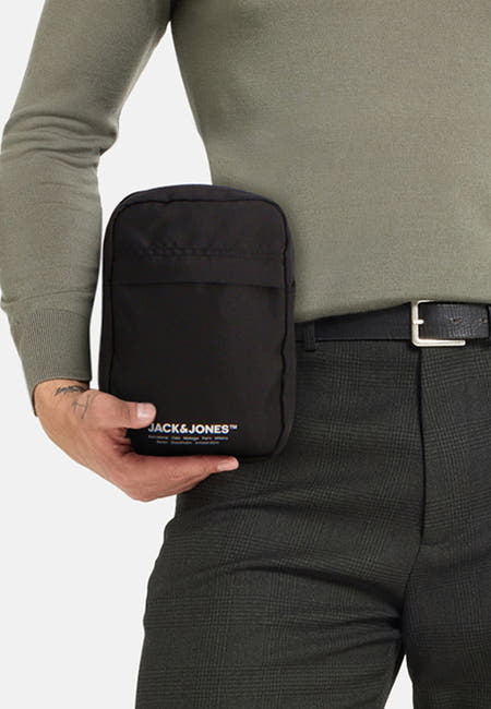 Sac à dos noir de Jack & Jones, tenu par une personne portant un pull olive et des pantalons.