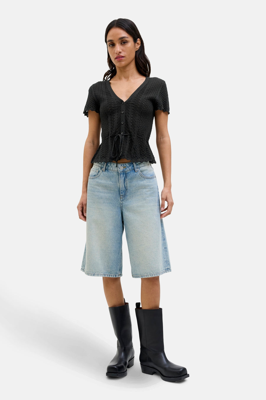 Pull noir JJXX avec fermeture à boutons et un détail noué à la taille, porté avec des shorts en denim et des bottes noires.