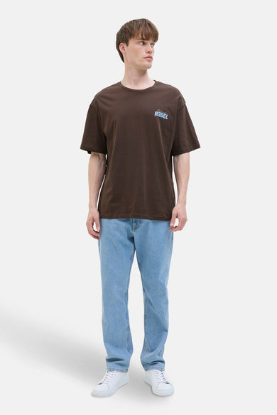 T-shirt brun à manches courtes de JJREBEL, avec une impression bleu clair en haut, porté avec un jean léger et des baskets blanches.