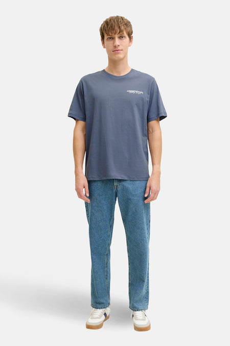Man met blauw T-shirt, jeans en sneakers, staand voor witte achtergrond.
