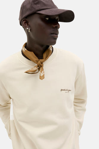 Gilet écru d'ORIGINALS BY JACK & JONES, avec un petit logo de marque et porté avec une bandana marron et un chapeau.