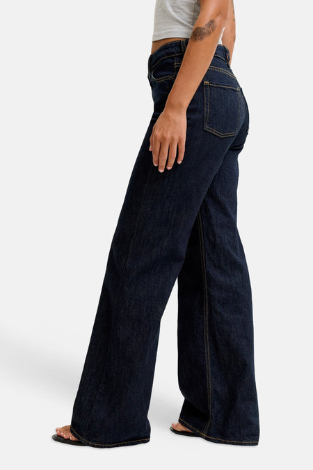 Jean évasé bleu foncé en denim de JJXX, profil latéral avec coutures jaunes et jambes larges, le modèle porte un crop top gris et des sandales noires