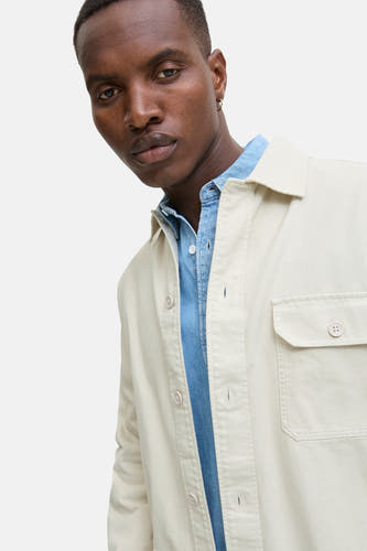 Chemise beige de PREMIUM BLUE par JACK & JONES, avec poche poitrine et fermeture à boutons, portée sur une chemise denim bleue.