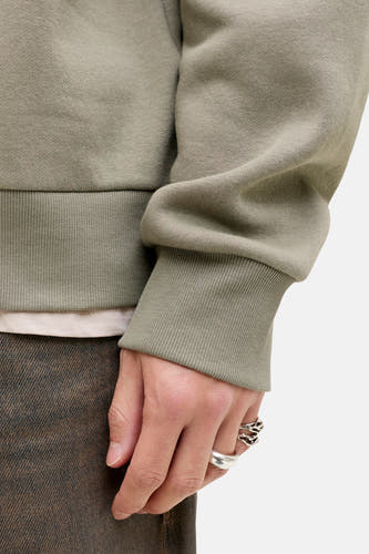 Pull-over vert de ORIGINALS BY JACK & JONES, avec des bordures côtelées larges, porté avec un pantalon marron et des bagues argentées.