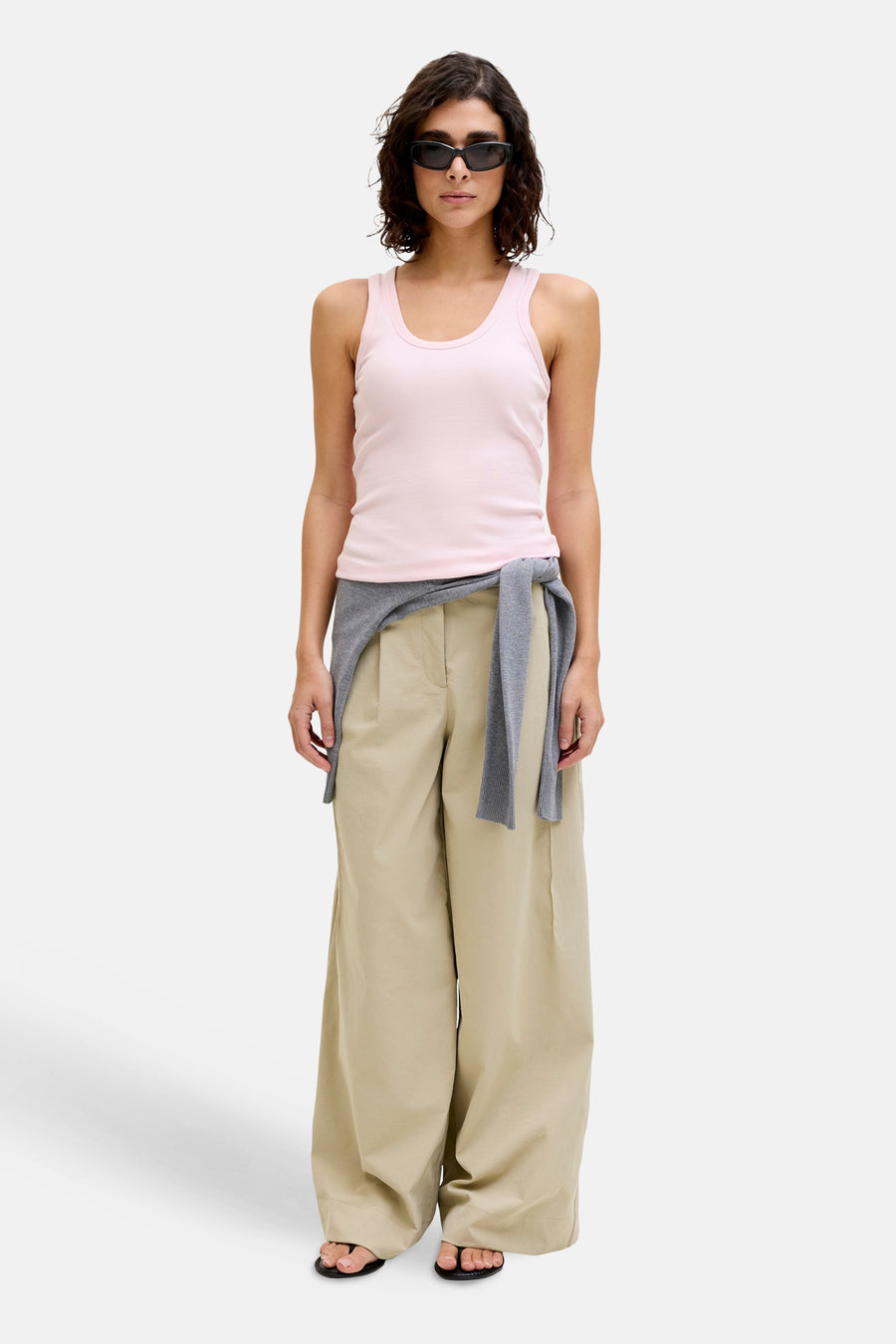 Débardeur côtelé rose de JJXX, stylé avec des lunettes de soleil, un pull gris noué à la taille et un pantalon beige ample.